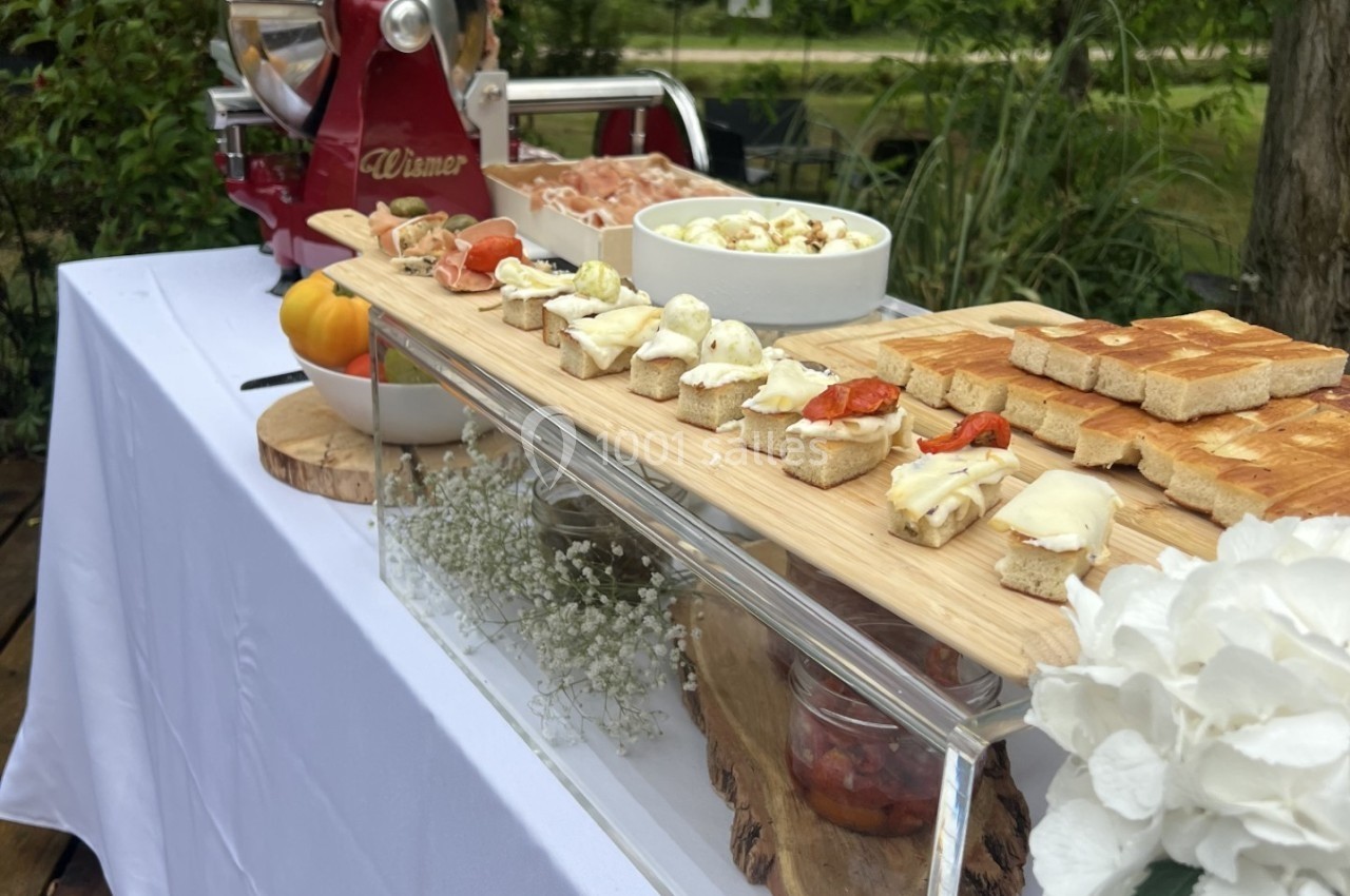 Plateau de toasts et amuse-bouches disposés sur une table blanche en extérieur, avec une trancheuse et un décor végétal.
