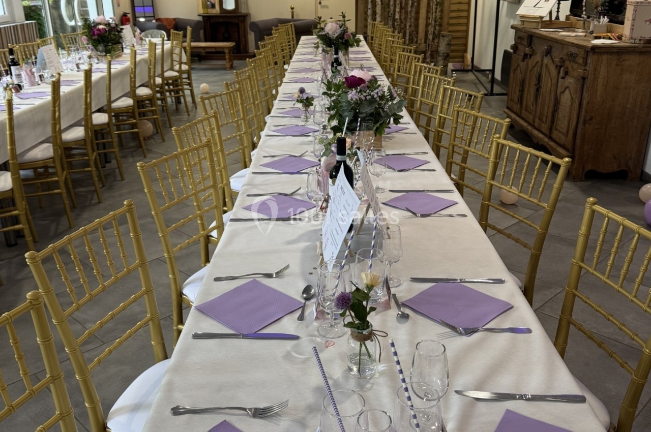 Grande table de réception décorée avec nappes blanches, serviettes violettes, fleurs et chaises dorées dans une salle…