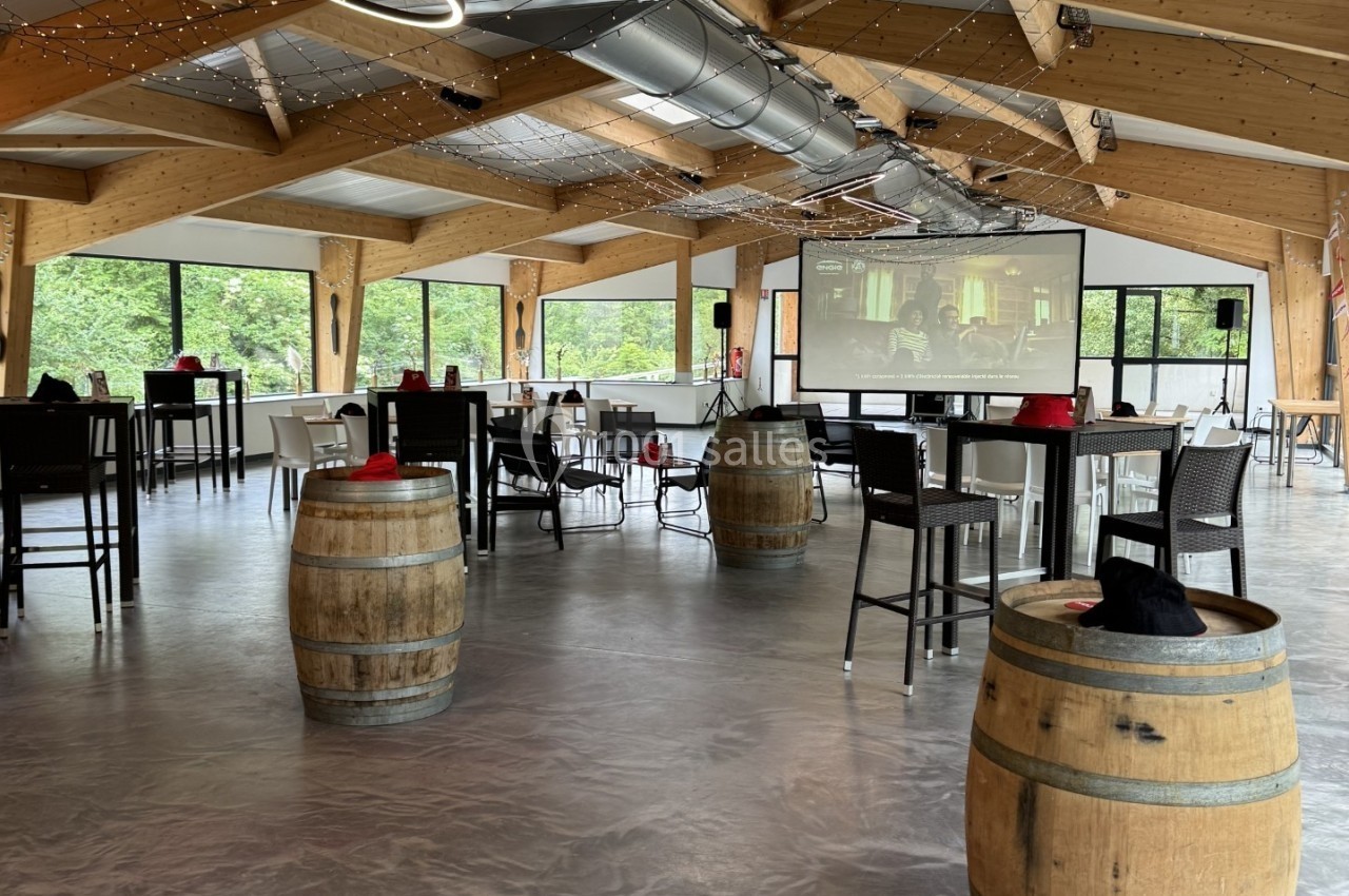 Salle lumineuse avec charpente en bois, tables dressées, tonneaux décoratifs et grandes baies vitrées donnant sur la verdure.