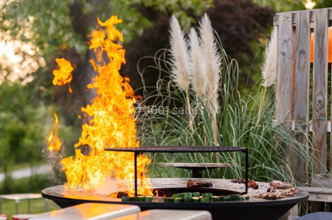 Barbecue en plein air avec flammes vives, grille de cuisson et végétation en arrière-plan.