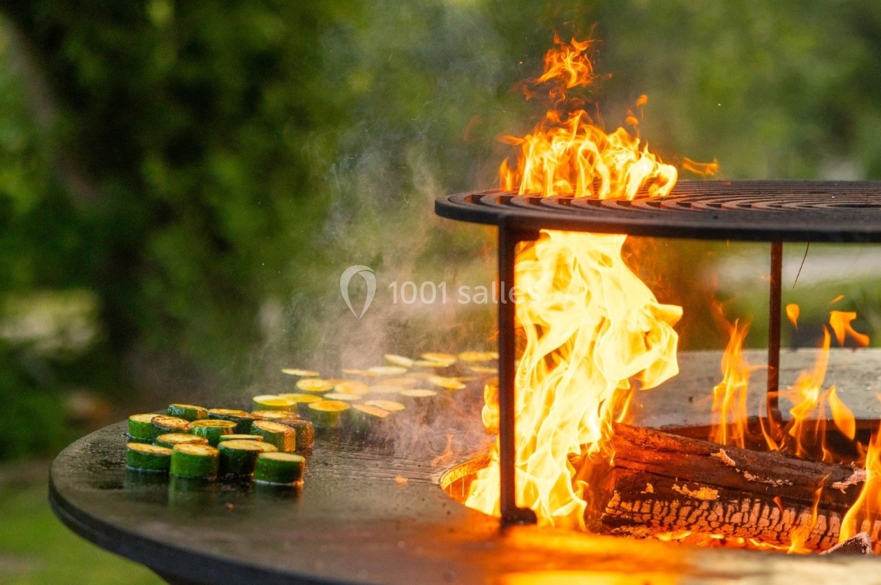 Légumes grillant sur un barbecue circulaire avec flammes vives en extérieur, entouré de verdure.