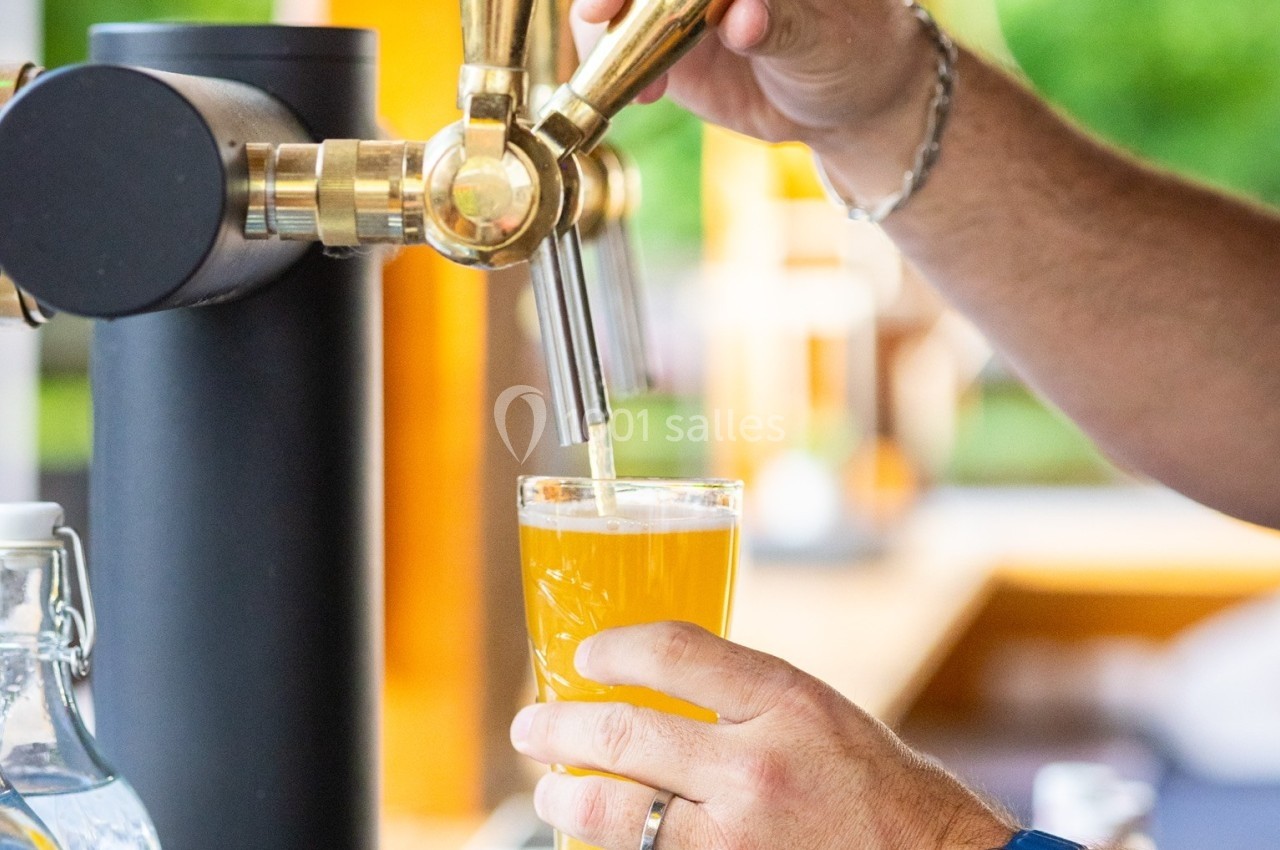 Une main sert une bière blonde à la pression dans un verre, avec un bar en arrière-plan.