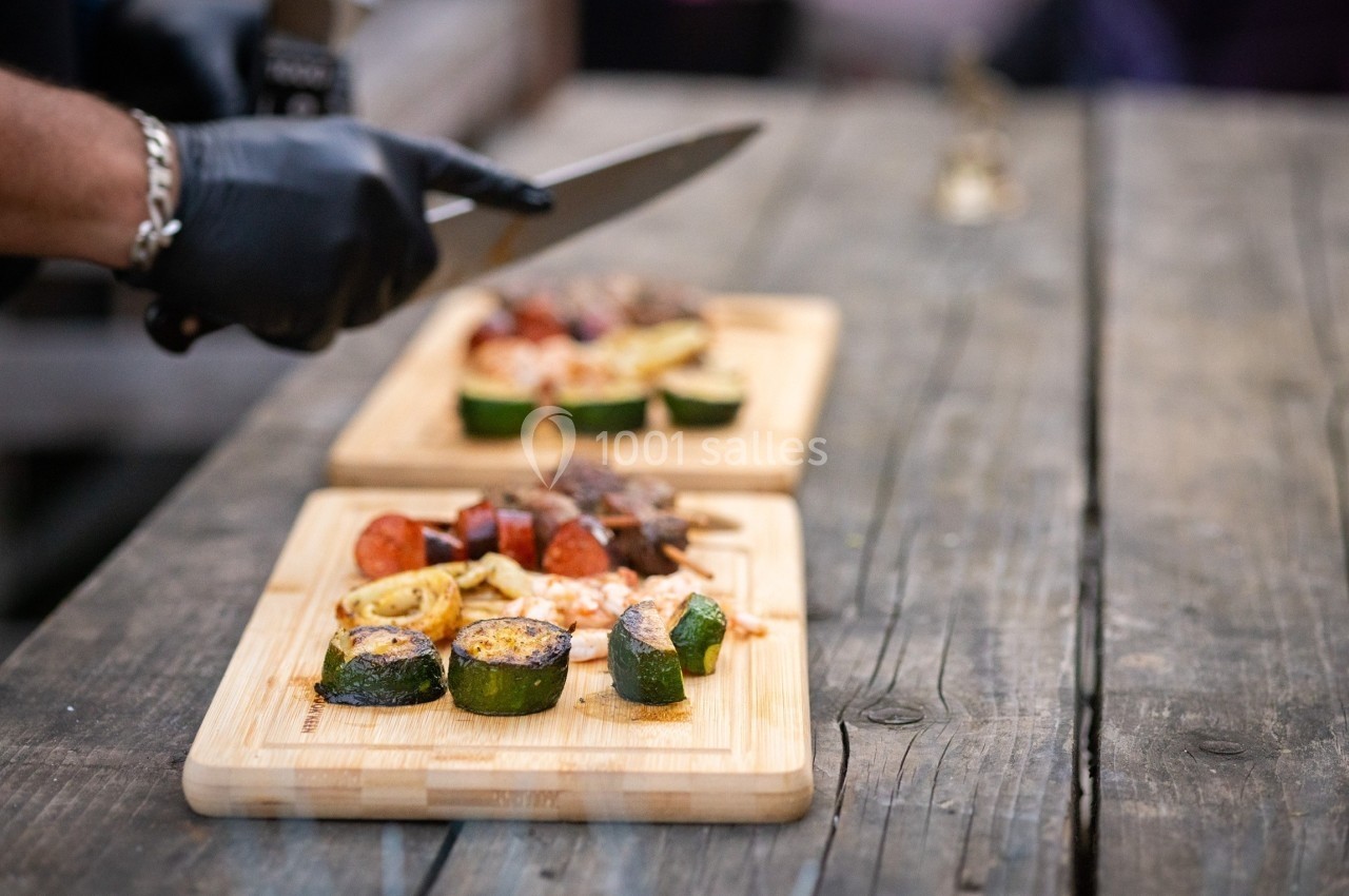 Un cuisinier découpe des aliments sur une planche en bois, avec des légumes grillés et de la viande en arrière-plan.