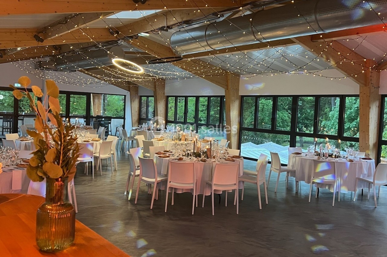 Salle de réception lumineuse avec tables décorées, grandes baies vitrées et plafond en bois orné de guirlandes lumineuses.