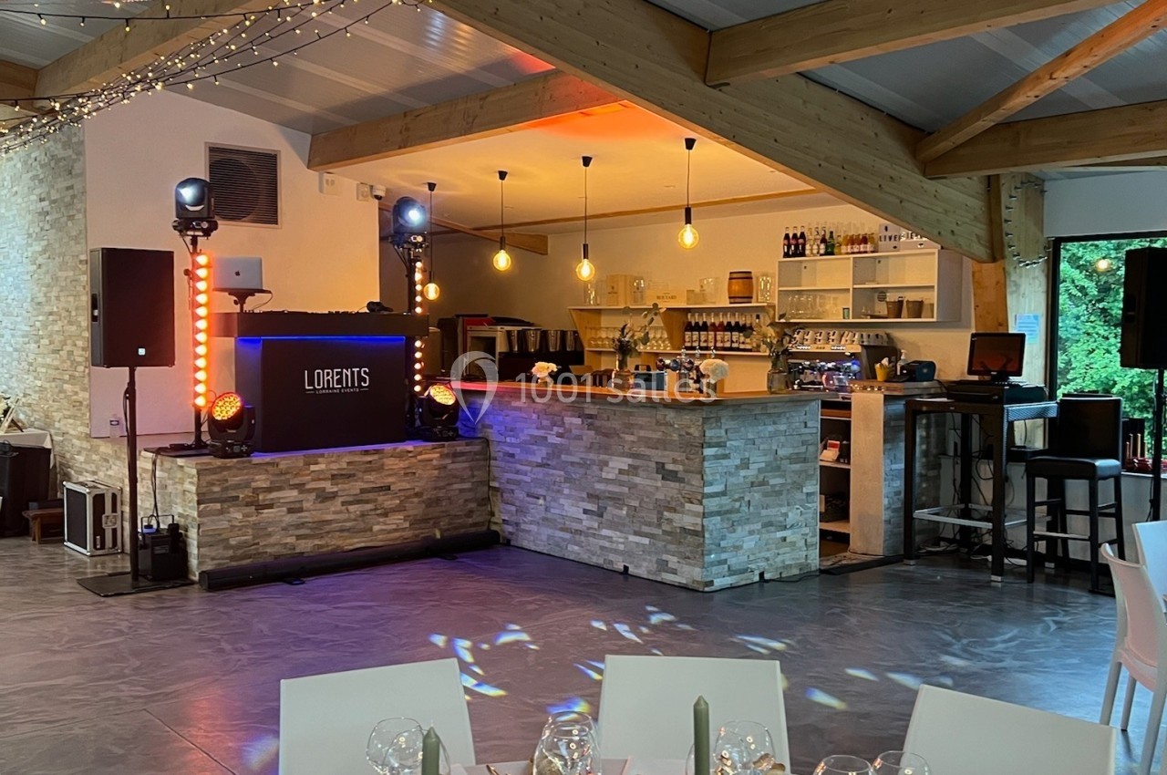 Salle de réception décorée avec des tables dressées, un bar en pierre, des guirlandes lumineuses et un espace DJ.