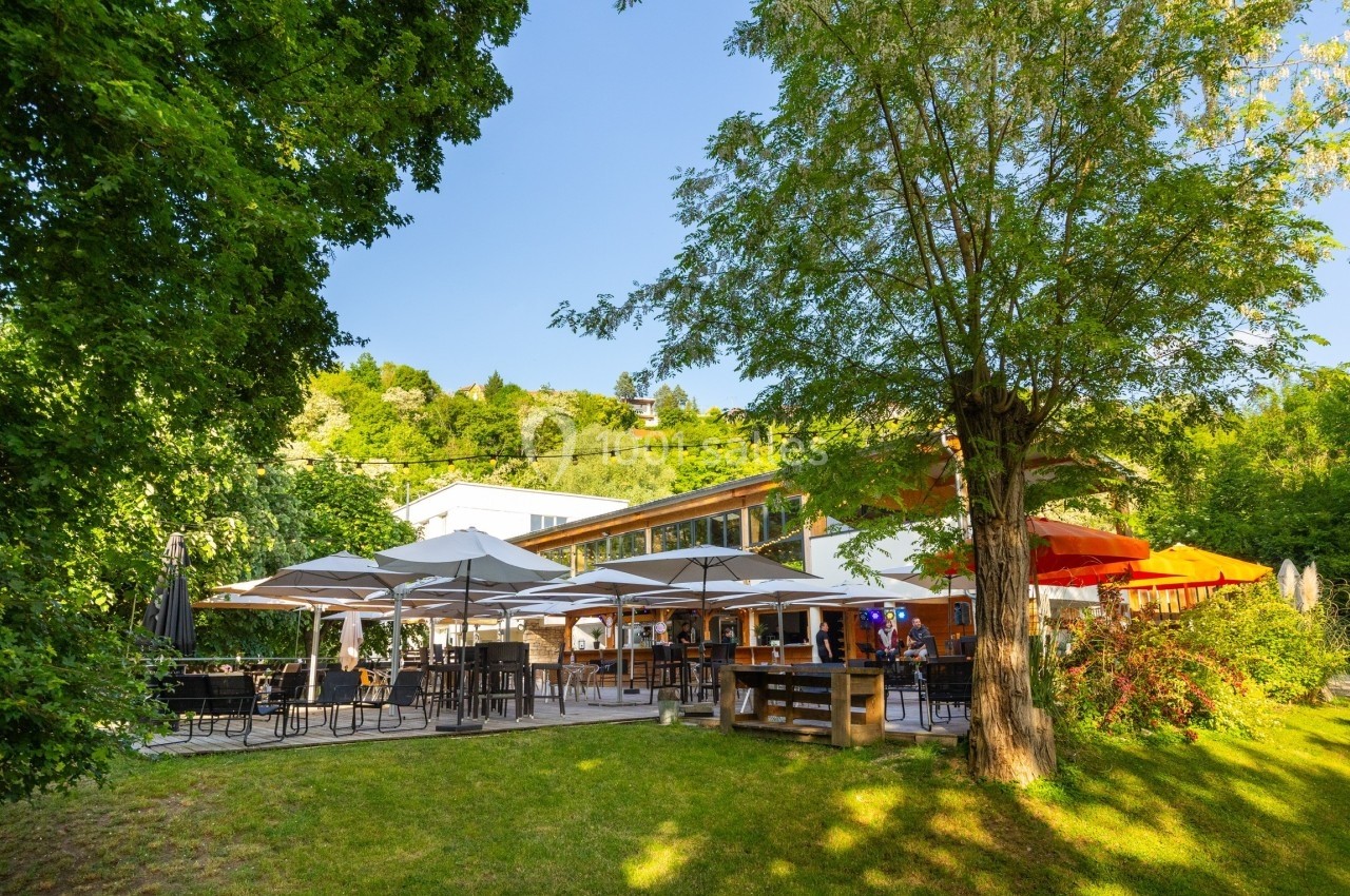 Terrasse d'un restaurant entourée de verdure, avec des parasols blancs et colorés sous un ciel dégagé.