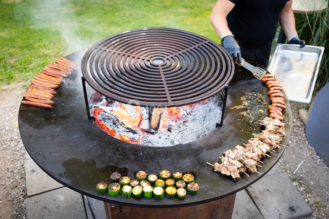 Barbecue circulaire avec saucisses, brochettes et légumes grillés, un cuisinier en arrière-plan tenant une plaque.