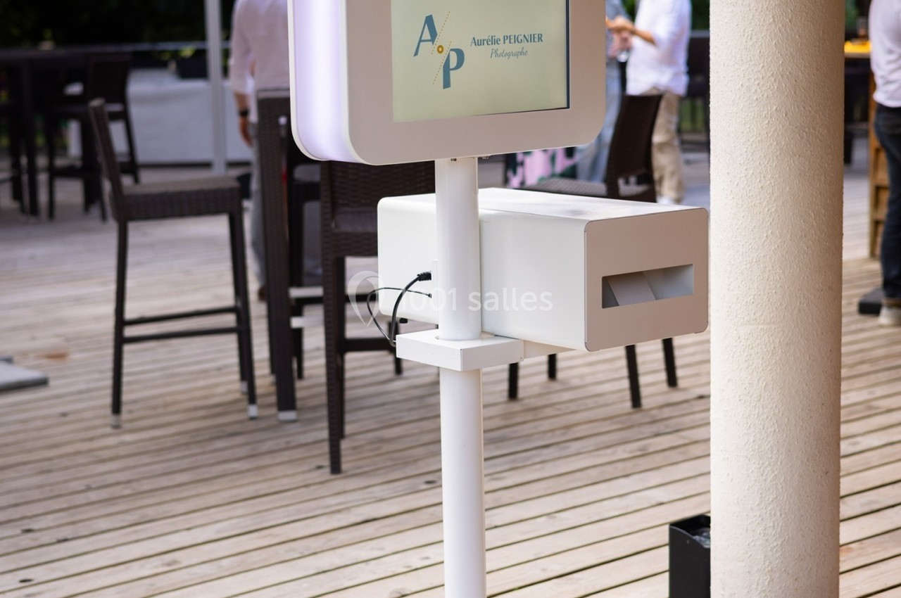 Borne photo interactive blanche installée sur une terrasse en bois, entourée de parasols et de tables hautes.