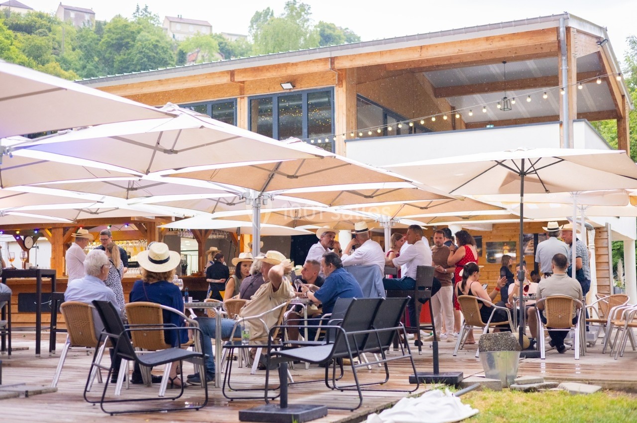 Personnes assises et debout sous des parasols sur une terrasse en bois devant un bâtiment moderne en bois.