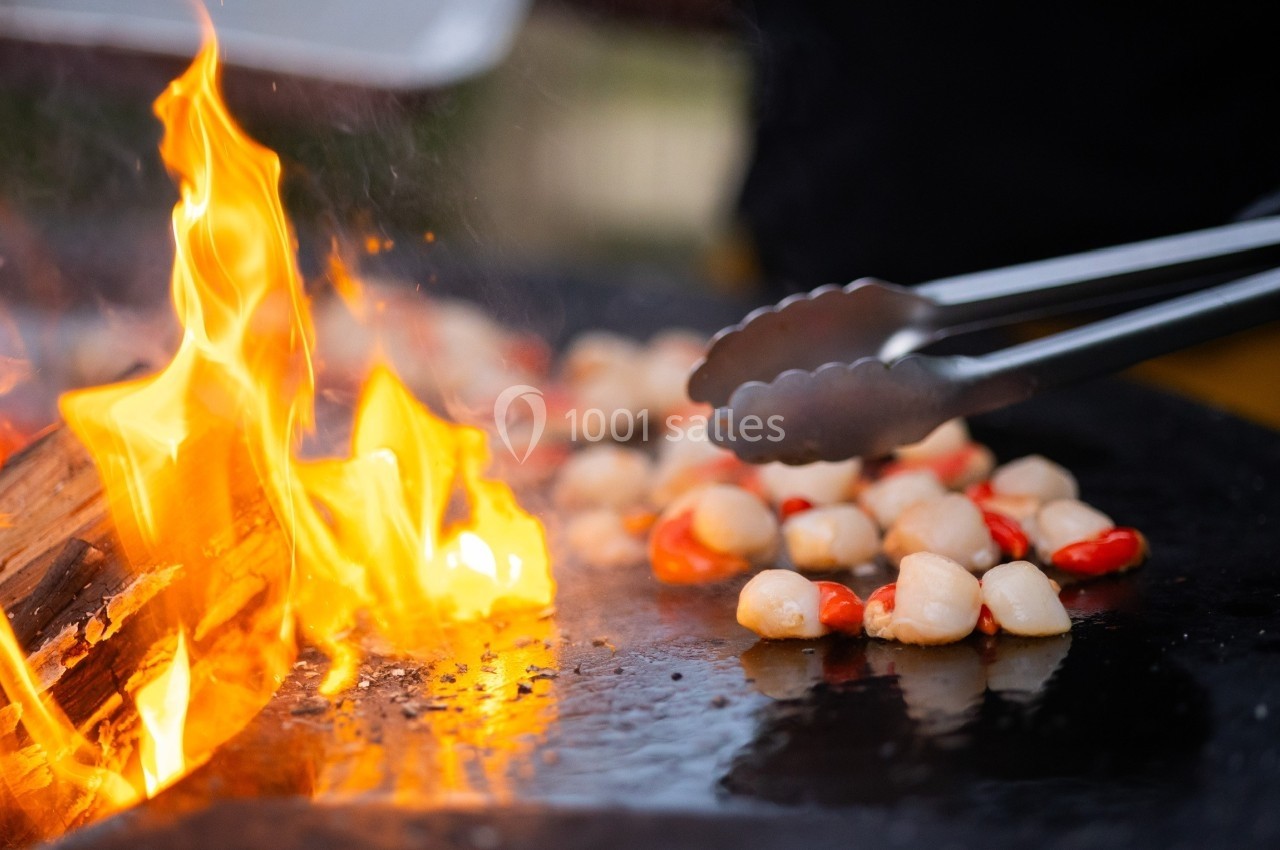 Des pinces retournent des noix de Saint-Jacques et des poivrons rouges sur une plaque chauffante près d'un feu vif.