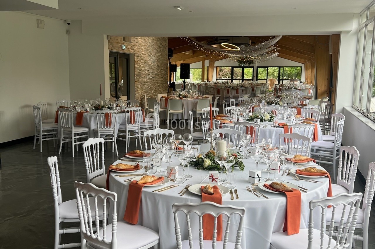 Salle de réception décorée avec des tables rondes, nappes blanches, serviettes orange et chaises blanches.