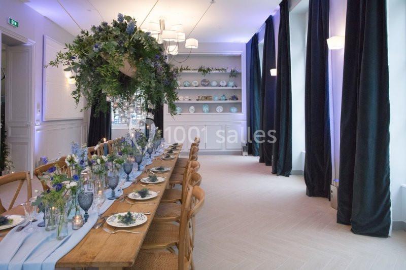 Grande table en bois décorée de fleurs bleues et vertes, dans une salle lumineuse avec rideaux sombres et étagères en…