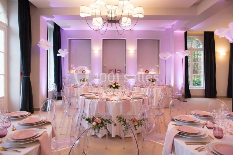 Salle de réception élégante décorée de tables rondes avec nappes blanches, fleurs roses et éclairage tamisé.