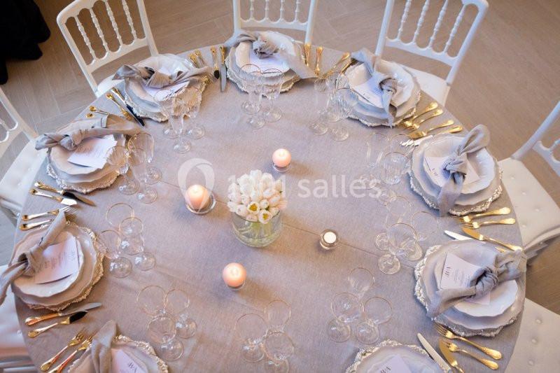 Table ronde élégamment dressée avec nappes grises, vaisselle raffinée, bougies et bouquet central de fleurs blanches.
