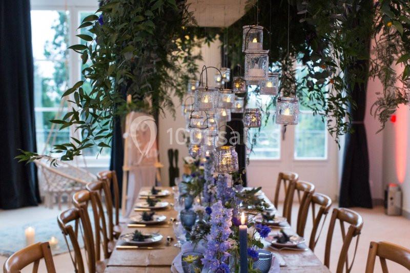 Table en bois décorée avec des fleurs violettes, des bougies suspendues et des feuillages, dans une salle lumineuse.