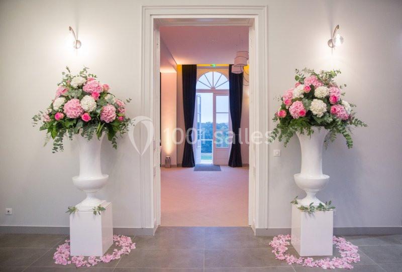 Deux grands vases blancs avec des bouquets de fleurs roses et blanches encadrent une porte ouverte sur une salle lumineuse.