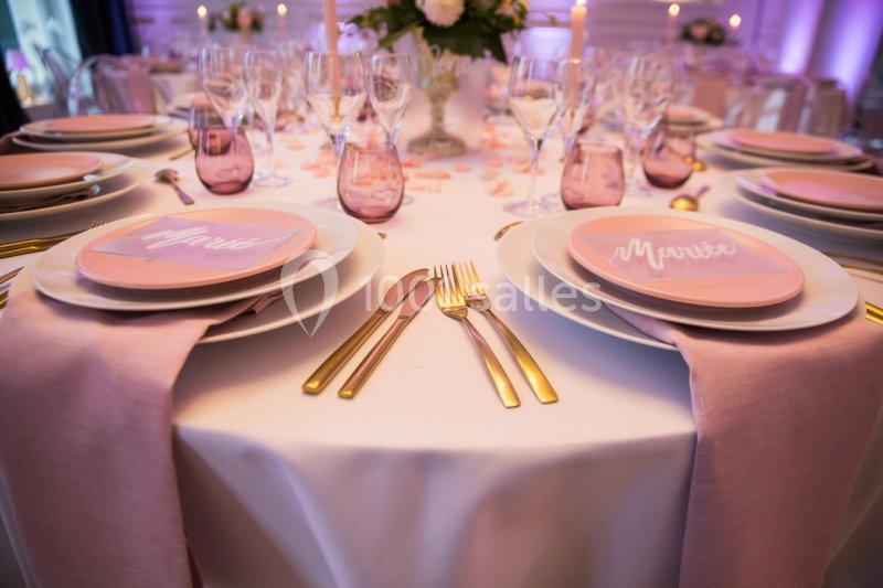 Table élégamment dressée avec nappes blanches, serviettes roses, couverts dorés et assiettes personnalisées.