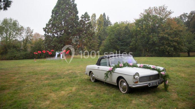 Voiture ancienne blanche décorée de fleurs, stationnée dans un parc verdoyant avec des arbres et des sculptures en arrière…