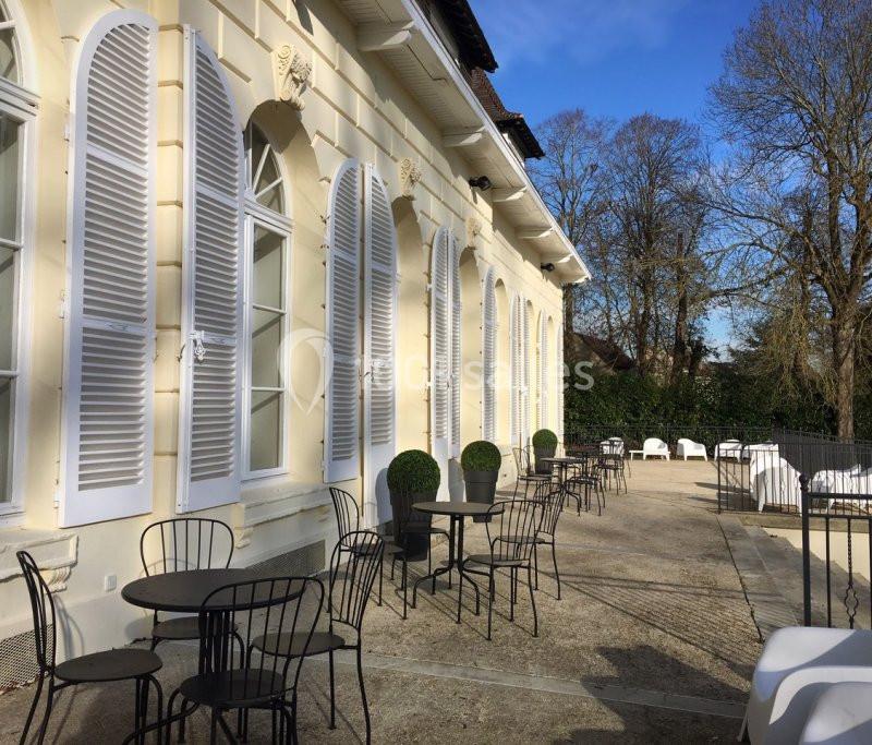 Terrasse ensoleillée avec tables et chaises en métal noir devant une façade beige aux volets blancs.
