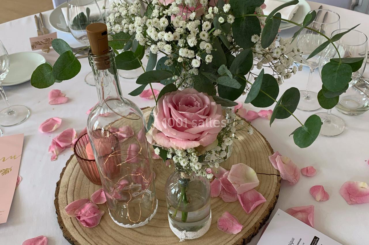 Centre de table avec roses roses, gypsophile, eucalyptus, bouteilles en verre et pétales sur une rondelle de bois.
