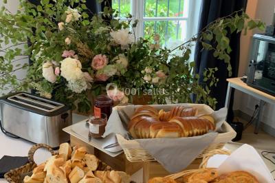 Buffet de petit-déjeuner avec viennoiseries, pain, confiture et grille-pain, devant une fenêtre et des fleurs décoratives.