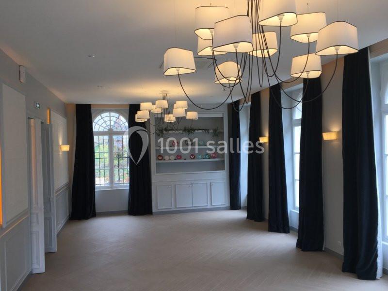 Salle lumineuse avec parquet clair, murs blancs, rideaux sombres, étagères décoratives et lustres modernes suspendus.