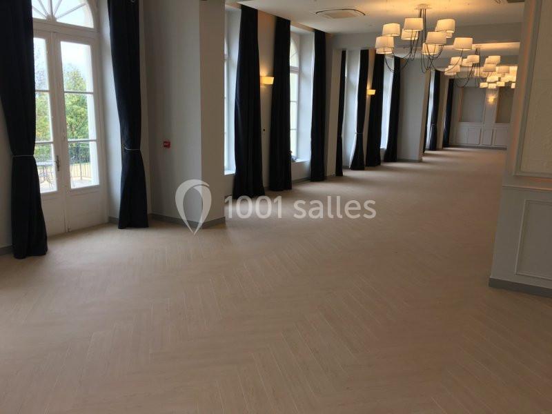 Salle lumineuse avec parquet clair, grandes fenêtres, rideaux noirs et lustres suspendus.