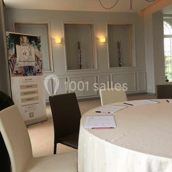 Salle lumineuse avec table ronde dressée, chaises, bloc-notes, et un panneau d'information en arrière-plan.