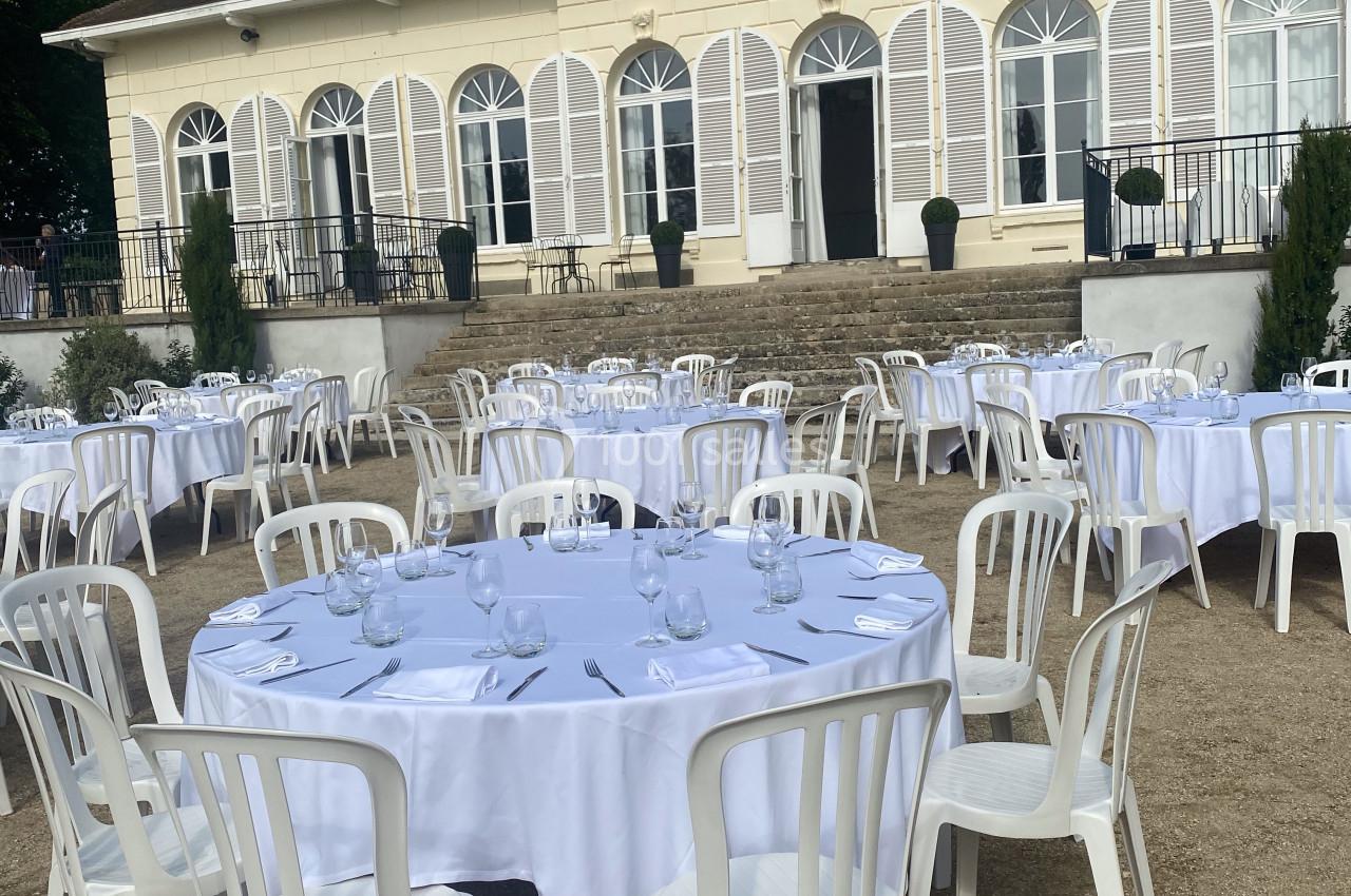 Tables rondes dressées avec nappes blanches et chaises en plastique devant une grande maison de style classique.