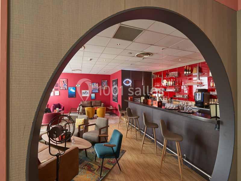 Vue d'un bar moderne avec comptoir, tabourets hauts, fauteuils colorés et décorations murales dans un espace chaleureux.