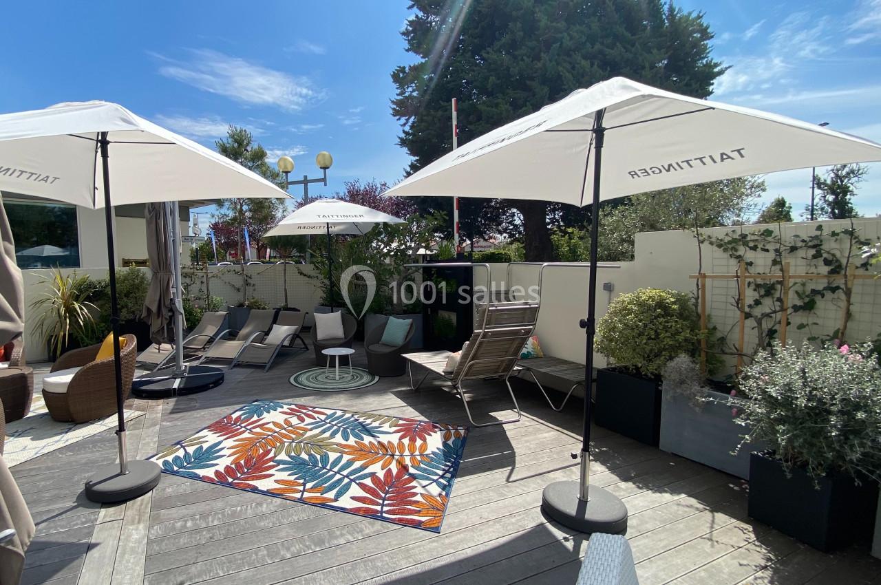 Terrasse ensoleillée avec parasols, chaises longues, tapis coloré et plantes en pots, entourée de verdure.