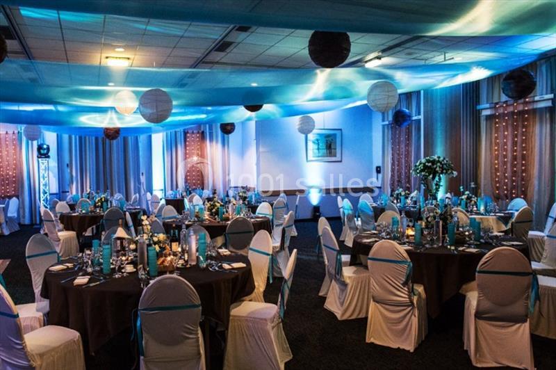 Salle de réception décorée avec des tables rondes, nappes blanches, centres de table et éclairage bleu tamisé.