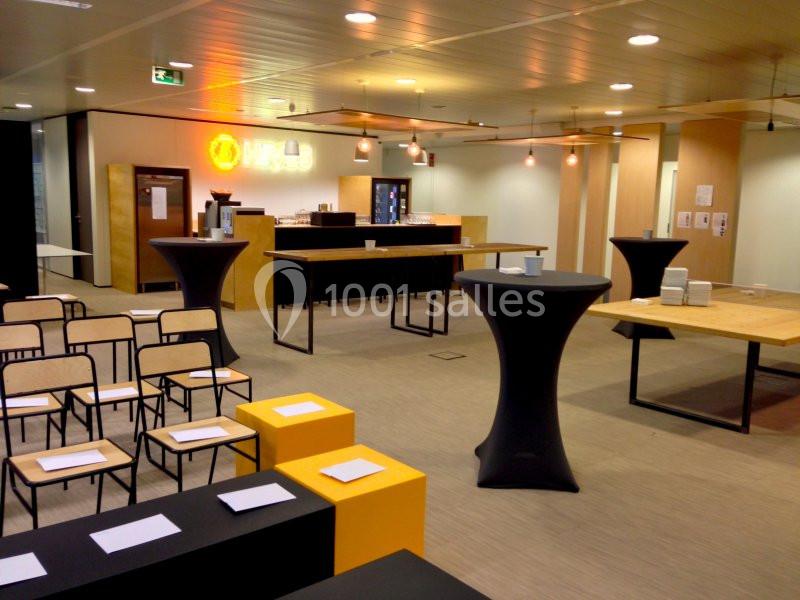 Salle de réunion avec chaises alignées, tables hautes noires et espace de restauration en arrière-plan.