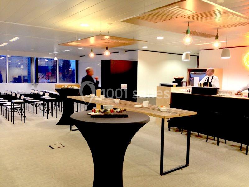 Salle lumineuse aménagée pour un événement, avec chaises alignées, tables hautes et un bar en arrière-plan.