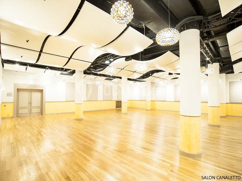 Salle spacieuse avec parquet en bois clair, colonnes blanches, plafonds ondulés et luminaires suspendus.