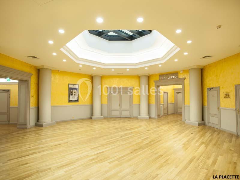 Salle lumineuse avec parquet en bois clair, murs jaunes, colonnes grises et plafond avec verrière octogonale.