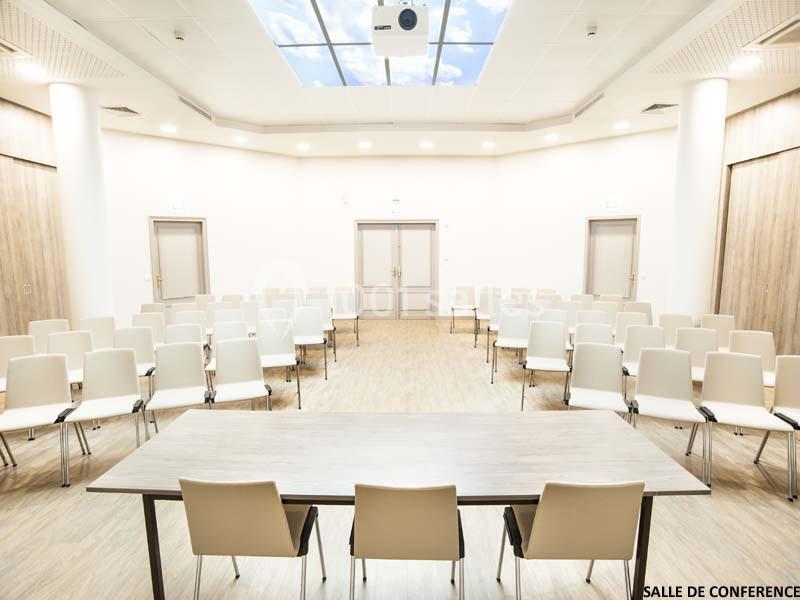Salle de conférence lumineuse avec des rangées de chaises blanches et une table en bois au premier plan.