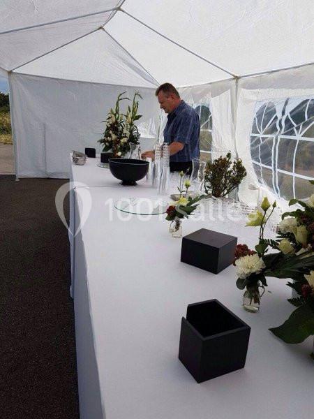 Un homme prépare une table décorée de fleurs sous une tente blanche, avec des verres et des accessoires disposés.