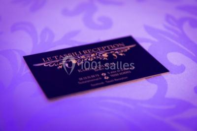 Salle de réception décorée avec des lumières violettes, une arche florale et des chaises transparentes disposées autour de…