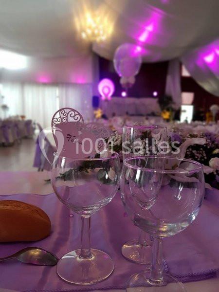 Trois verres à pied sur une table décorée de fleurs et de nappes violettes dans une salle de réception éclairée.