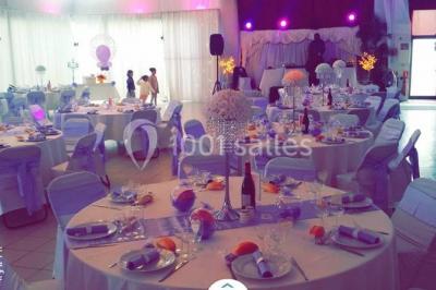 Salle de réception décorée avec des lumières violettes, une arche florale et des chaises transparentes disposées autour de…