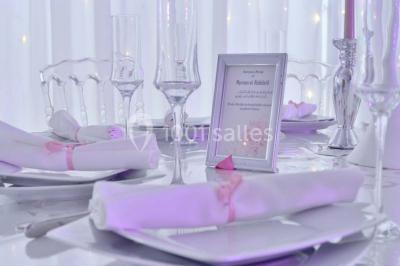 Salle de réception décorée avec des lumières violettes, une arche florale et des chaises transparentes disposées autour de…