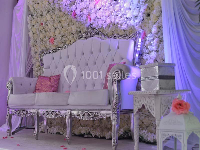 Canapé blanc capitonné avec accoudoirs sculptés, devant un mur de fleurs blanches et des décorations roses.