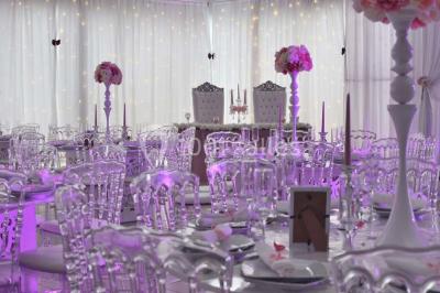 Salle de réception décorée avec des lumières violettes, une arche florale et des chaises transparentes disposées autour de…