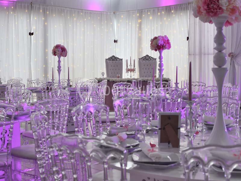 Salle de réception décorée avec des tables transparentes, des fleurs roses et un éclairage violet tamisé.