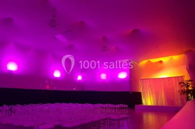 Salle de réception décorée avec des lumières violettes, une arche florale et des chaises transparentes disposées autour de…