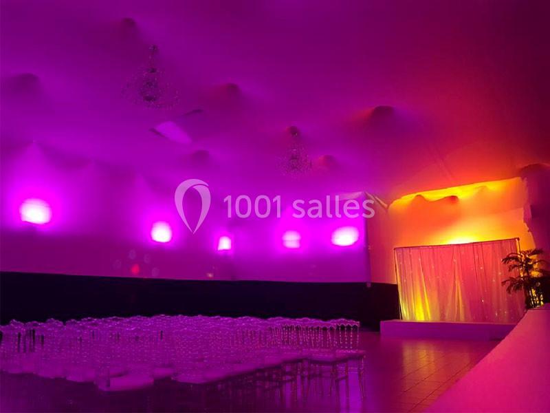 Salle de réception éclairée par des lumières violettes et jaunes, avec des chaises transparentes alignées devant une scène.