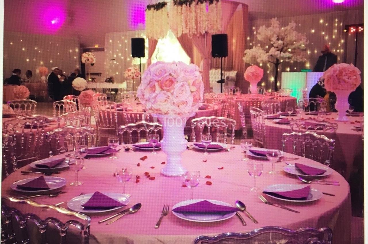 Salle décorée pour un événement avec tables rondes dressées, nappes roses, centres de table fleuris et éclairage tamisé.