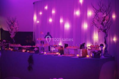 Salle de réception décorée avec des lumières violettes, une arche florale et des chaises transparentes disposées autour de…