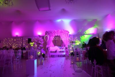 Salle de réception décorée avec des lumières violettes, une arche florale et des chaises transparentes disposées autour de…